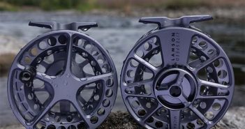 Two fly reels sitting on rocks next to a river.