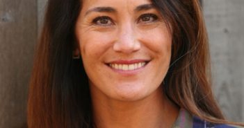 A woman in a plaid shirt smiling in front of a wooden wall.