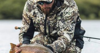 A man in a camouflage jacket is holding a brown trout.