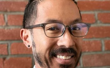A man in glasses smiles in front of a brick wall.