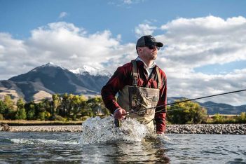 A man is fly fishing in a river with mountains in the background.