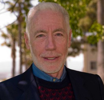 A man with white hair and a sweater in front of a tree.