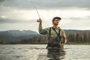 A man is fly fishing in a river with mountains in the background.