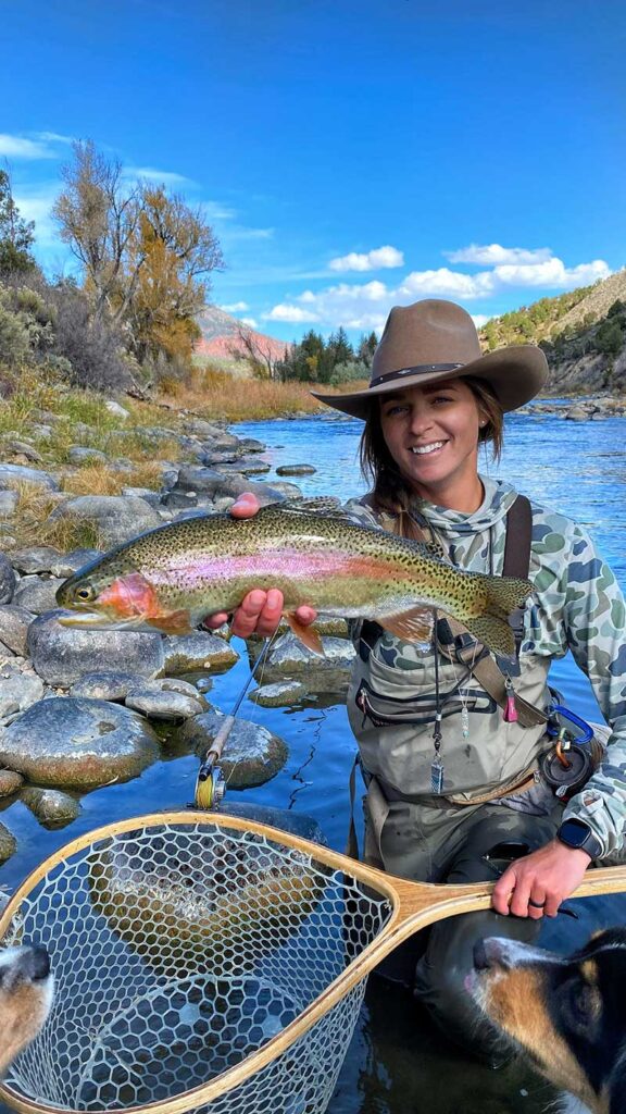 A woman holding a rainbow trout with her dog.