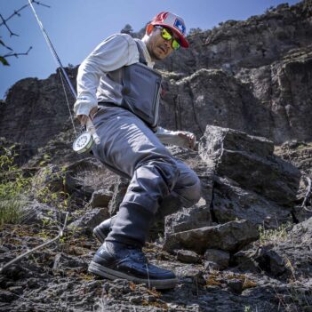 A fisherman wearing waders stepping over rocks with a fishing rod in hand.