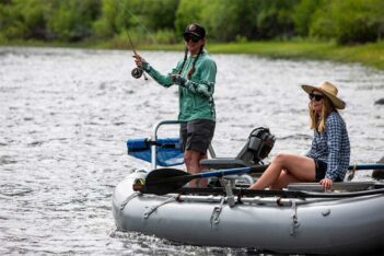 Two individuals fishing from an inflatable boat on a river, one standing and casting a line, the other seated at the stern.