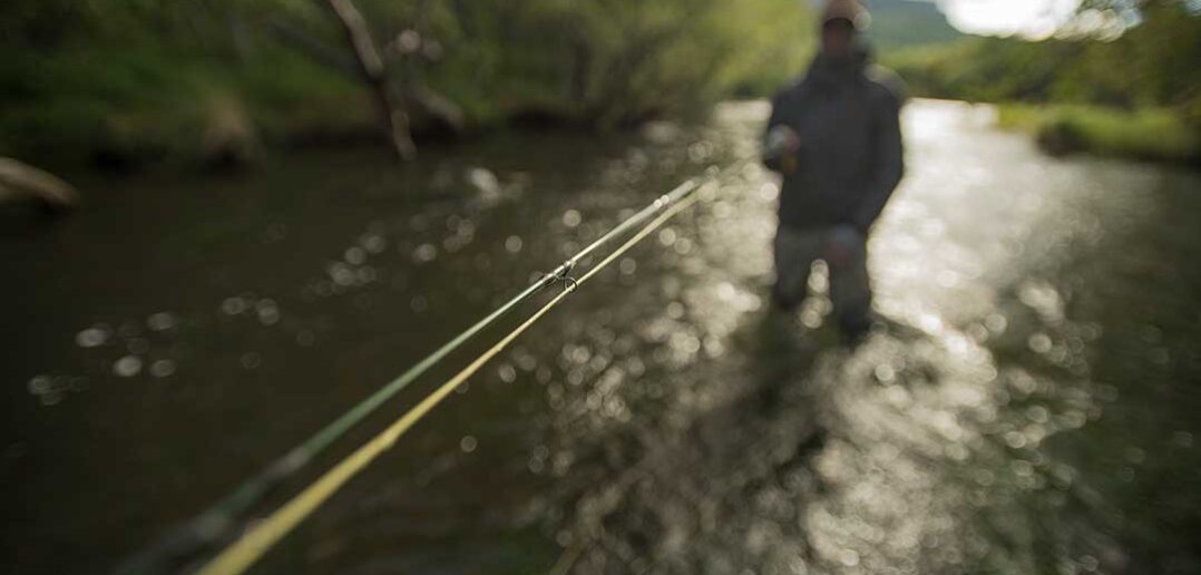 A person wading in a stream with a focus on a fishing rod in the foreground and the angler slightly blurred in the background.