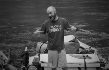 Man with a beard making hand gestures while standing on a boat.