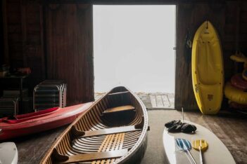 A variety of watercraft and gear stored in a wooden boathouse with an open door leading to a bright exterior.