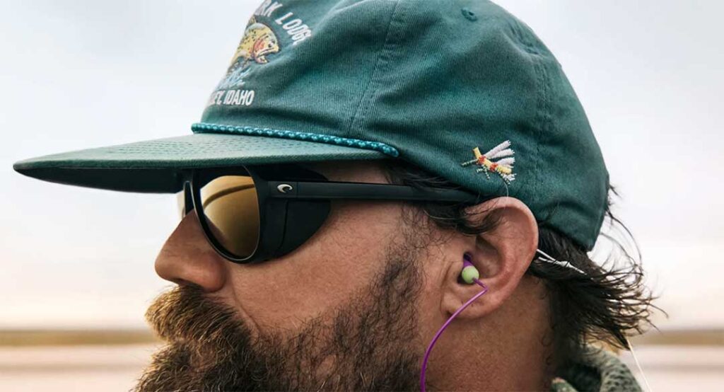 Close-up of a man wearing sunglasses and a green cap with pins, featuring a side profile with a focus on his earbud.