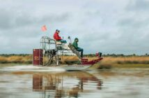Two people ride on an airboat across a water body with grassy shores, one sitting and the other standing, both wearing jackets and caps.