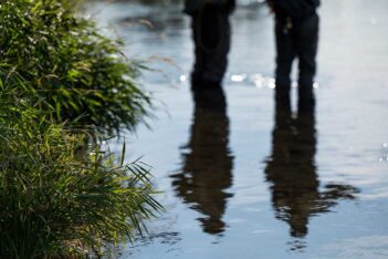 Silhouettes of two people with fishing gear reflected in the water near green grass plants at the water's edge.