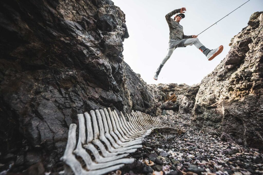 A person jumps across a rocky gap above an animal skeleton lying on the ground among rocks and shells.