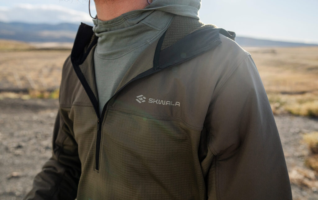 A person outdoors wears a green Skwala jacket with a hood and quarter-zip, standing in a sunlit field with mountains in the background.