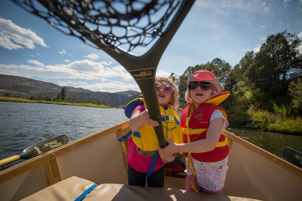Two young children in life jackets and sunglasses sit in a canoe on a river, holding up a fishing net towards the camera with trees and hills in the background.