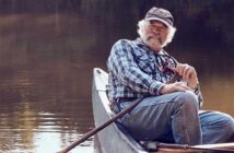 An older man with a white beard sits in a canoe on calm water, holding a fishing rod and wearing a plaid shirt, jeans, and a cap.