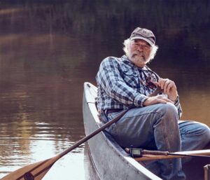 An older man with a white beard sits in a canoe on calm water, holding a fishing rod and wearing a plaid shirt, jeans, and a cap.