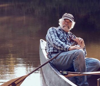 An older man with a white beard sits in a canoe on calm water, holding a fishing rod and wearing a plaid shirt, jeans, and a cap.