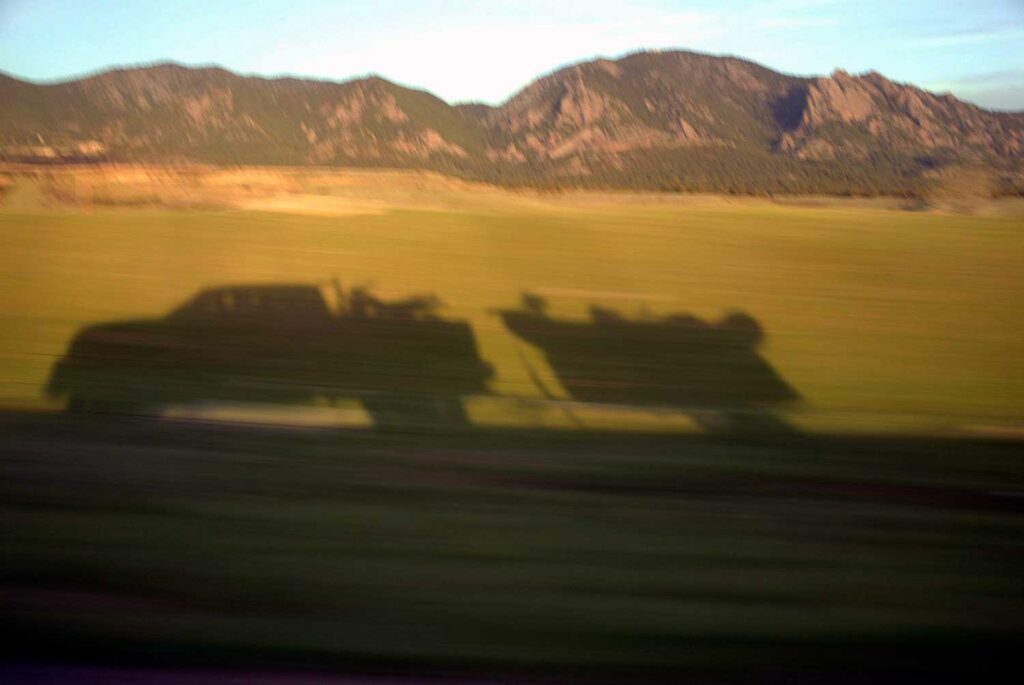 Shadow of a pickup truck towing a boat cast onto a grassy field, with mountains in the background.