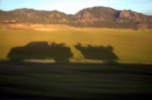 Shadow of a pickup truck towing a boat cast onto a grassy field, with mountains in the background.