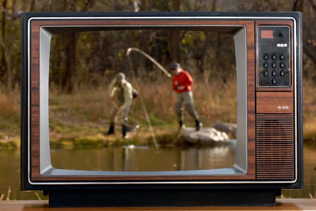 Vintage television displaying an image of two people fishing by a pond, with trees and grass visible in the background.