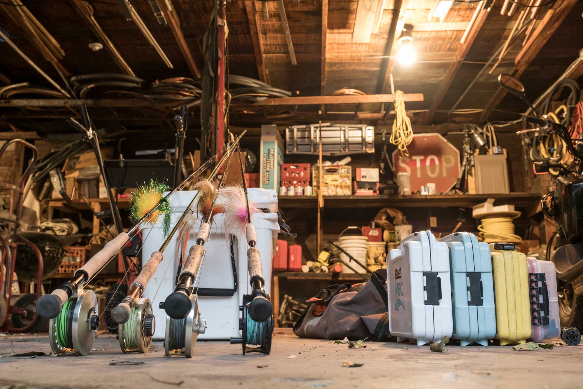 Several fishing rods rest against a cooler in a cluttered garage, with tackle boxes and various tools and equipment visible on the shelves in the background.