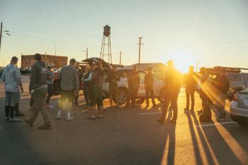 A group of people stand and talk in a parking lot near parked cars at sunrise or sunset, with long shadows on the ground.