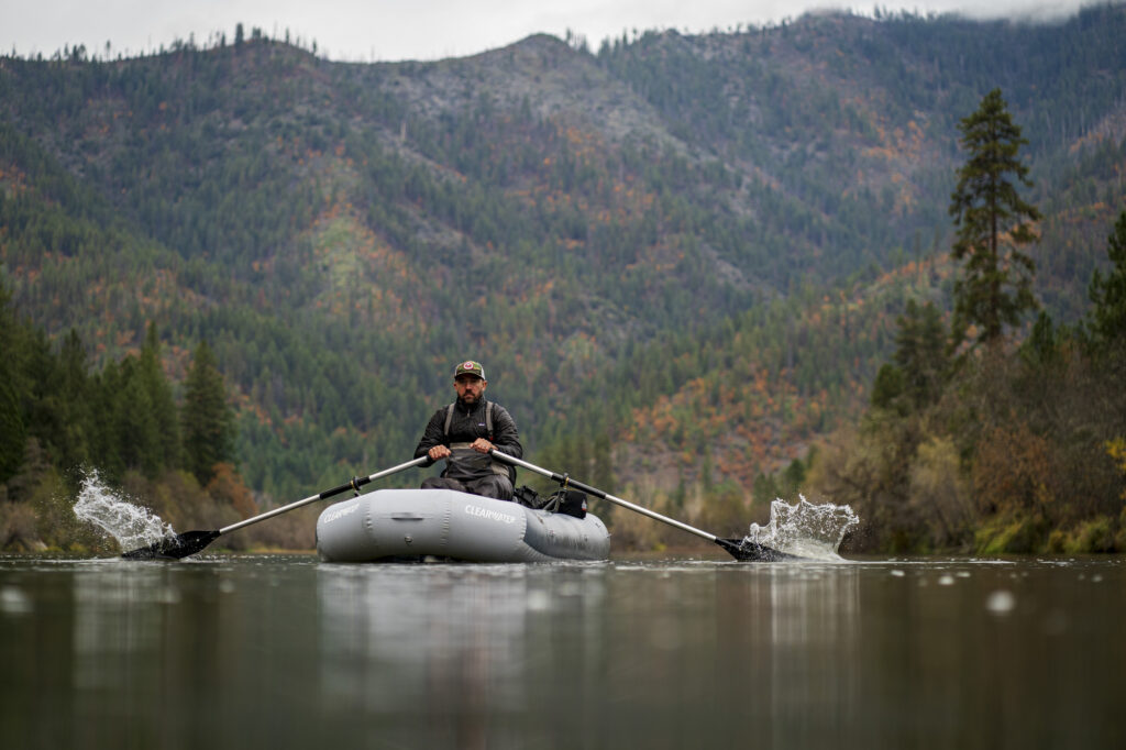 A man rows an inflatable raft on a calm river with forested mountains in the background under an overcast sky.