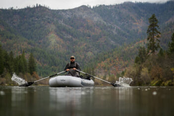A man rows an inflatable raft on a calm river with forested mountains in the background under an overcast sky.
