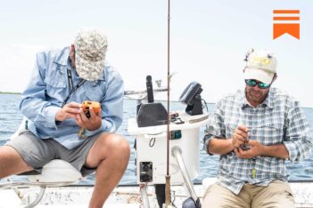 Two men sit on a boat checking devices, with fishing gear and electronics visible around them on a sunny day.