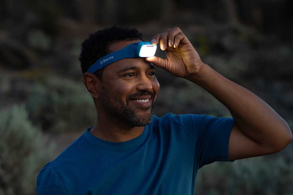 A man in a blue shirt adjusts a blue headlamp on his forehead, smiling outdoors in low light.