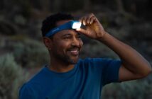 A man in a blue shirt adjusts a blue headlamp on his forehead, smiling outdoors in low light.