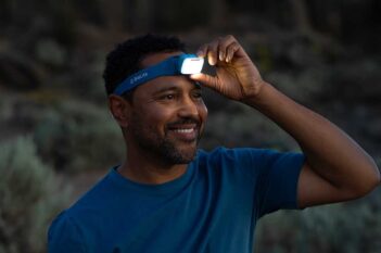 A man in a blue shirt adjusts a blue headlamp on his forehead, smiling outdoors in low light.