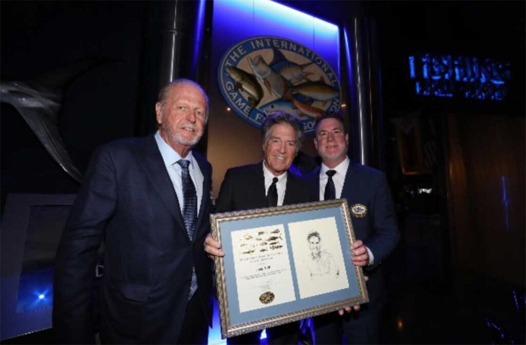 Three men in suits stand together at an event, with two of them holding a framed certificate and artwork beneath a large fishing-related logo.