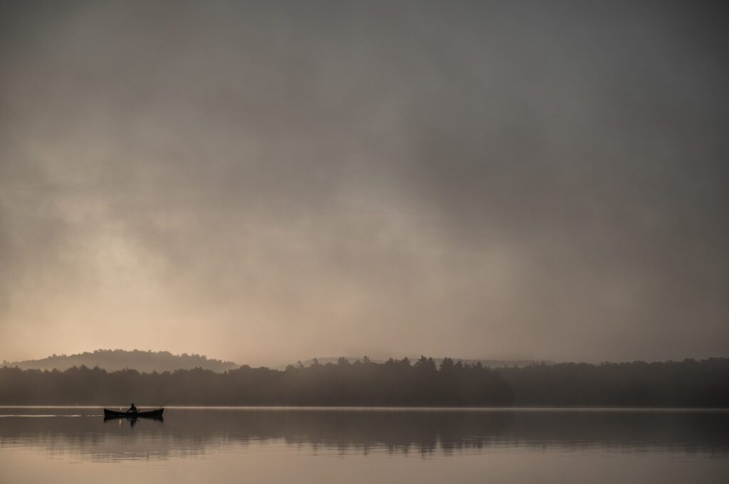 A small boat with two people floats on a calm lake at sunrise, surrounded by mist and distant tree-covered hills.