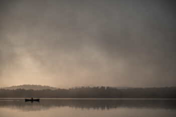 A small boat with two people floats on a calm lake at sunrise, surrounded by mist and distant tree-covered hills.