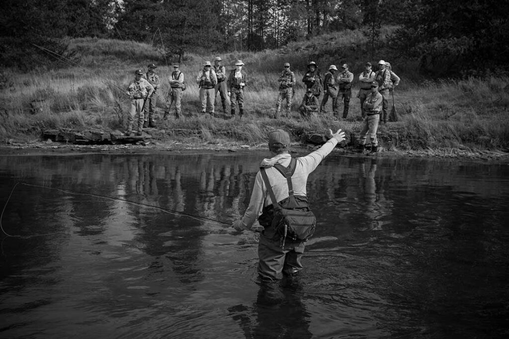 A person stands in a river fly fishing, gesturing toward a group of people watching from the grassy bank on the opposite side.
