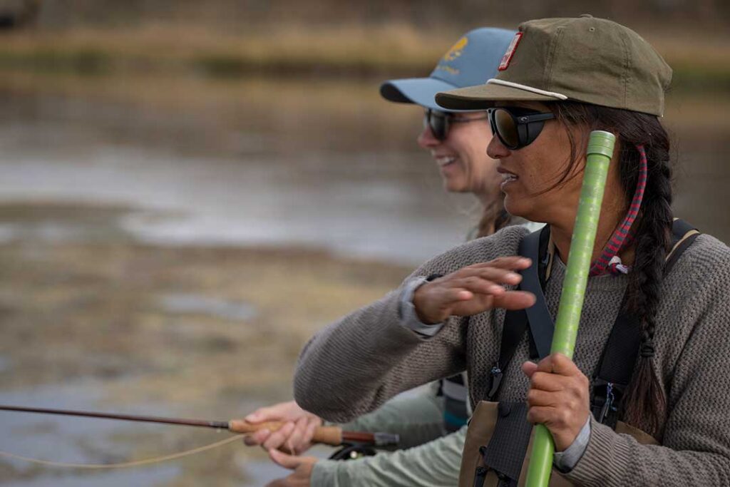 Two people wearing caps and sunglasses stand near a body of water, holding fishing rods and appearing to converse or focus on fishing.