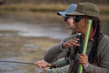 Two people wearing caps and sunglasses stand near a body of water, holding fishing rods and appearing to converse or focus on fishing.