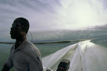 A person wearing sunglasses stands on a moving boat, with water and a cloudy sky in the background and the boat’s wake visible behind.