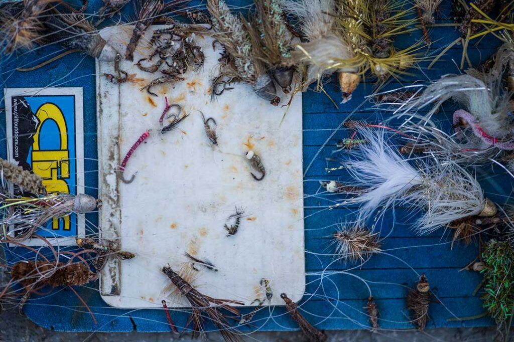 A variety of fishing flies and lures are arranged on a blue surface with a white, stained fly case and a partially visible sticker.