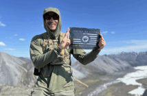 A person in outdoor gear holds a sign reading “United We Stand for Public Lands” on a mountain with rocky terrain and a river in the background.