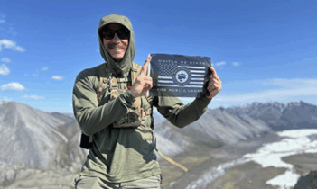 A person in outdoor gear holds a sign reading “United We Stand for Public Lands” on a mountain with rocky terrain and a river in the background.
