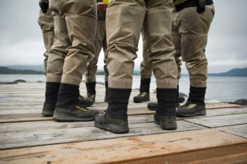 A group of people wearing khaki uniforms and black boots stand on a wooden dock near a body of water under a cloudy sky.