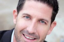 A man with short dark hair, light eyes, and a trimmed beard smiles at the camera, wearing a dark jacket and a light-colored shirt.