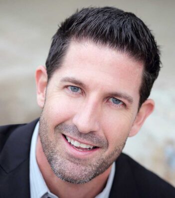 A man with short dark hair, light eyes, and a trimmed beard smiles at the camera, wearing a dark jacket and a light-colored shirt.