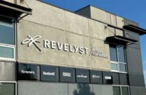 The exterior of a building displays the "Revelyst Outdoor Performance" sign along with logos for Simms, Bushnell, Primos, Camp Chef, and Sebile against a gray wall.