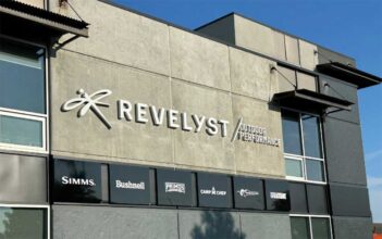 The exterior of a building displays the "Revelyst Outdoor Performance" sign along with logos for Simms, Bushnell, Primos, Camp Chef, and Sebile against a gray wall.