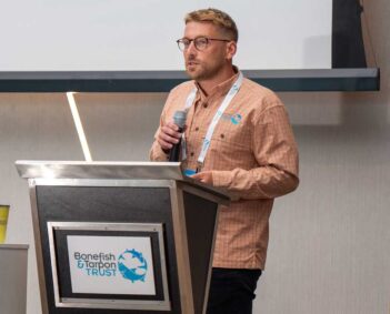 A man wearing glasses and a Bonefish & Tarpon Trust shirt speaks into a microphone at a podium with the organization’s logo during a presentation.