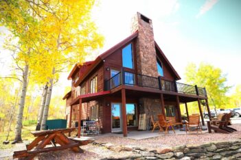 A two-story wooden cabin with a stone chimney, outdoor seating, and picnic table, surrounded by trees with yellow leaves on a sunny day.